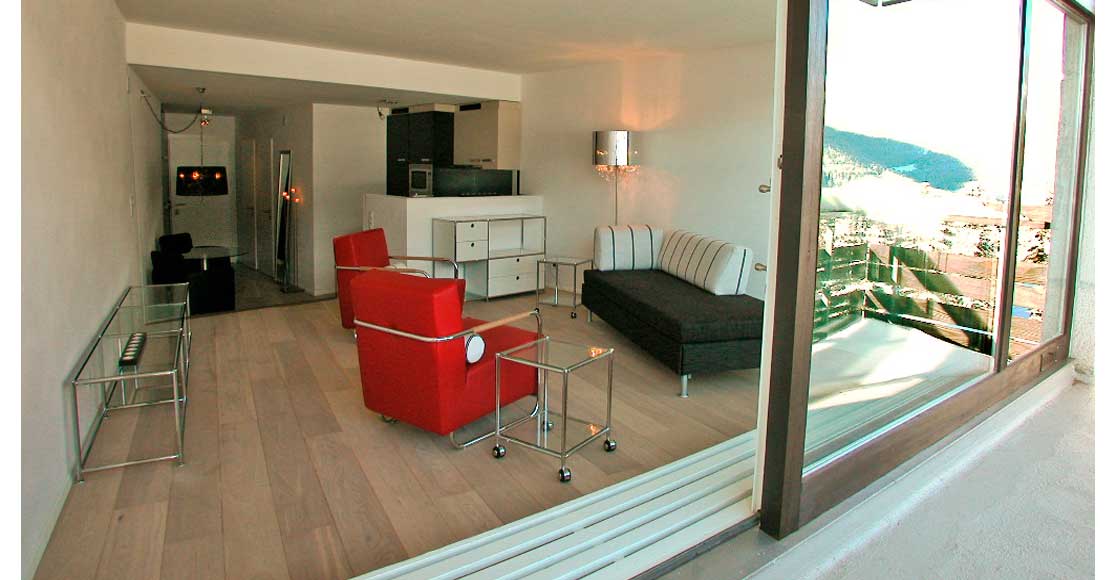Foto: Einblick vom Balkon in den Wohnraum mit roten Sesseln, Divan schwarz/weiss und Küche.